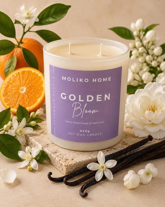 The Golden Bloom candle in its cream glass vessel with a lilac label, beside oranges, orange blossom and vanilla pods.